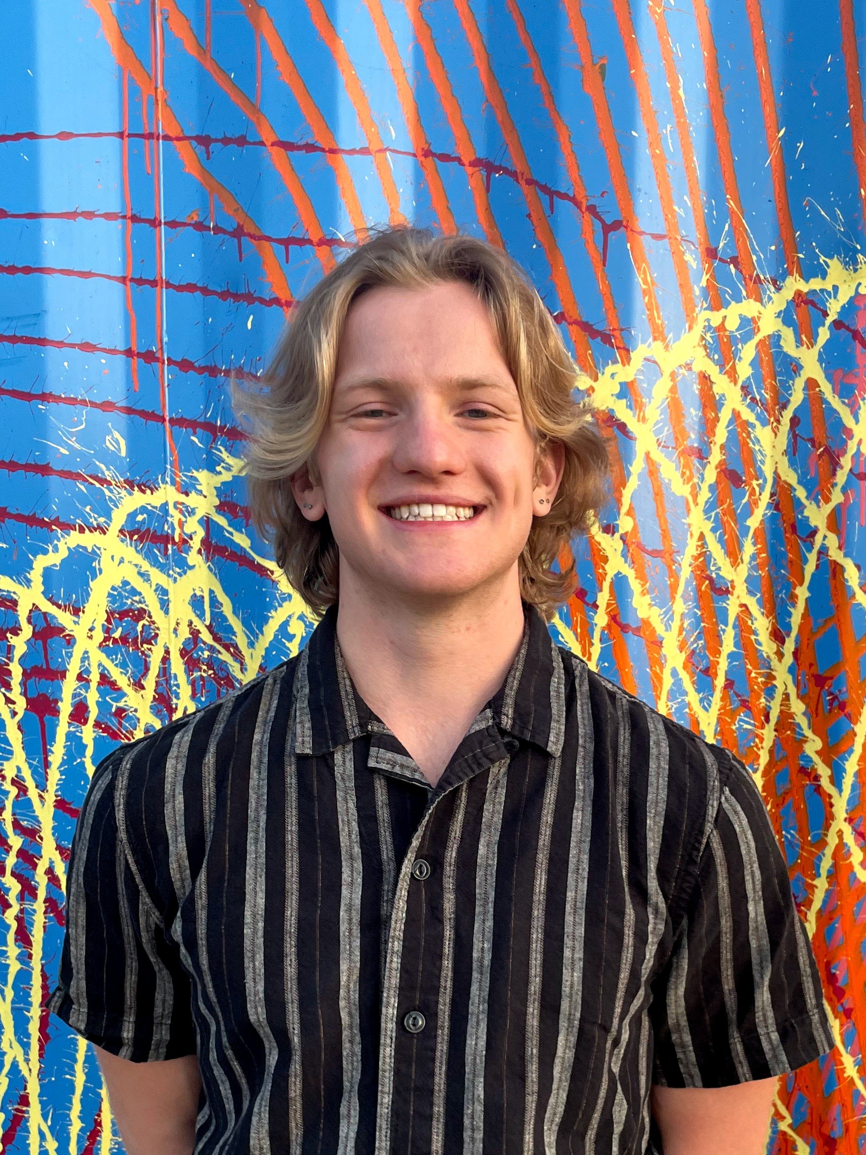 A man with light brown and a black striped shirt stands smiling behind a colorful wall