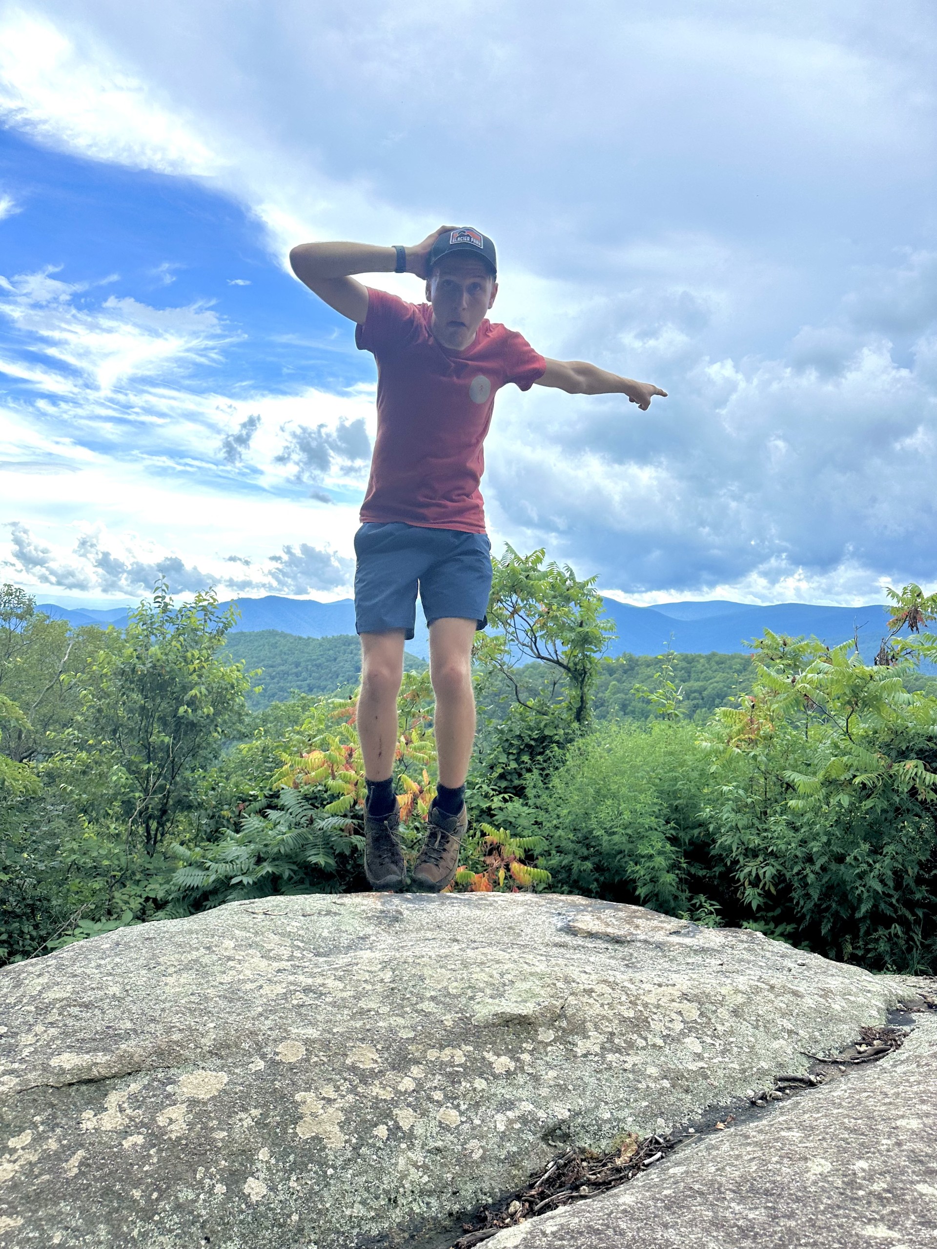 A man in a cap and red shirt standing on the edge of a rock pointing to the forest in the background