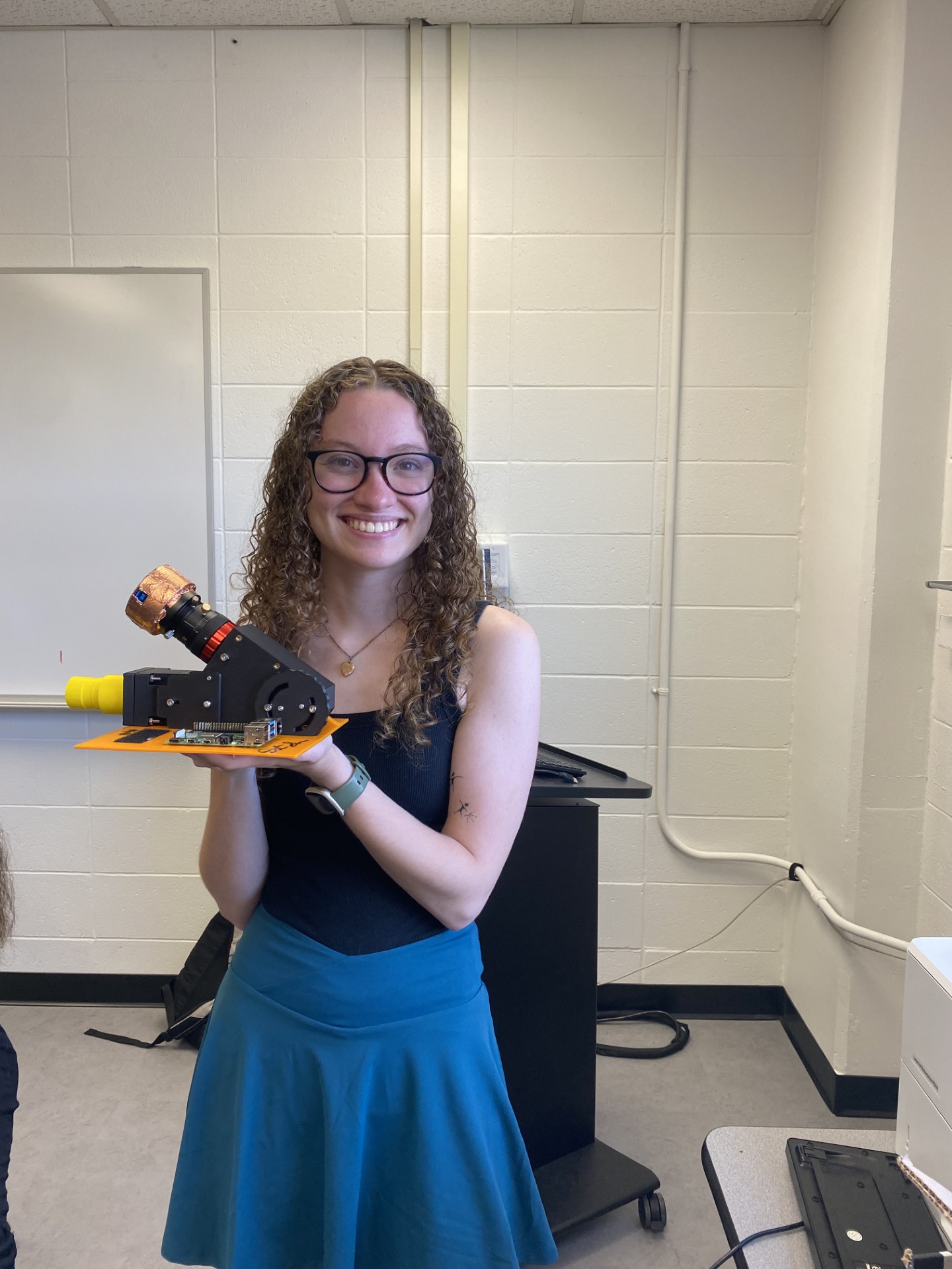 A woman with brown curly hair, glasses, and a blue dress holding electronics